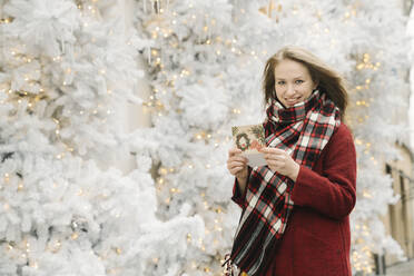 Porträt einer lächelnden jungen Frau, die mit einer Weihnachtskarte vor einem weißen Weihnachtsbaum auf der Straße steht - AHSF01842