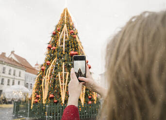 Crop-Ansicht einer Frau, die an einem verschneiten Tag den Weihnachtsbaum mit ihrem Smartphone fotografiert - AHSF01845