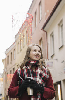 Porträt einer lächelnden jungen Frau mit Reisetasse zur Weihnachtszeit in der Stadt - AHSF01846