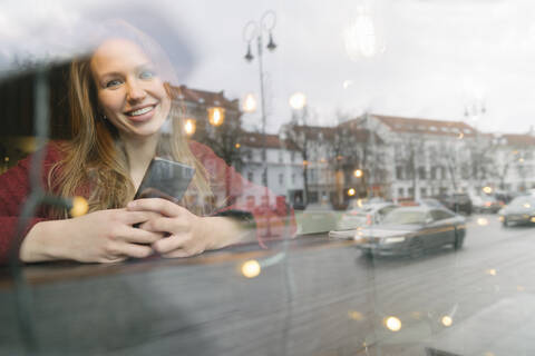 Porträt einer glücklichen jungen Frau mit Mobiltelefon in einem Café, die aus dem Fenster schaut, lizenzfreies Stockfoto