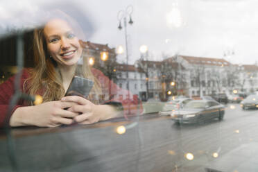 Portrait of happy young woman with mobile phone in a coffee shop looking out of window - AHSF01847