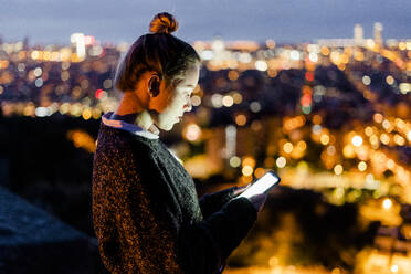 Junge Frau sitzt auf einem Geländer über der Stadt und benutzt ein Mobiltelefon, Barcelona, Spanien - GIOF07939