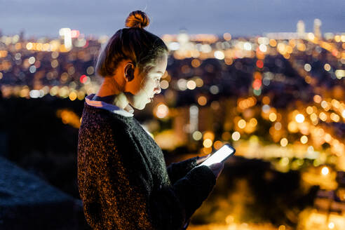 Junge Frau sitzt auf einem Geländer über der Stadt und benutzt ein Mobiltelefon, Barcelona, Spanien - GIOF07939