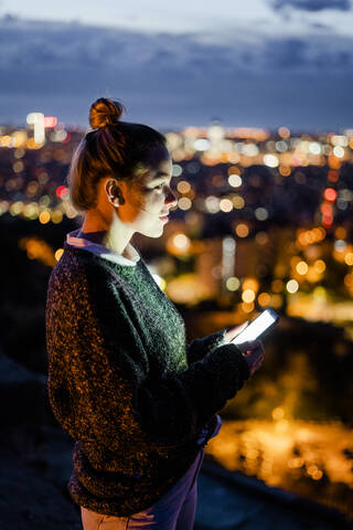 Junge Frau sitzt auf einem Geländer über der Stadt und benutzt ein Mobiltelefon, Barcelona, Spanien, lizenzfreies Stockfoto