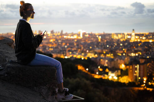 Junge Frau sitzt auf einem Geländer über der Stadt und benutzt ein Mobiltelefon, Barcelona, Spanien - GIOF07941