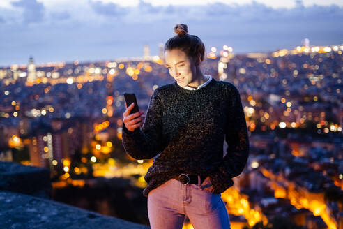 Junge Frau sitzt auf einem Geländer über der Stadt und benutzt ein Mobiltelefon, Barcelona, Spanien - GIOF07943