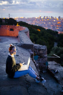 Junge Frau sitzt auf einem Geländer über der Stadt und benutzt ein Mobiltelefon, Barcelona, Spanien - GIOF07946