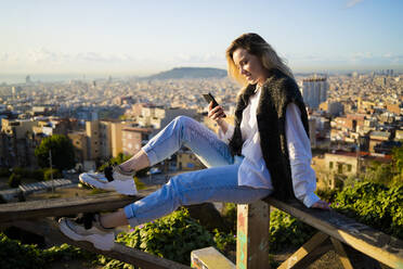 Young woman sitting on railing above the city using cell phone, Barcelona, Spain - GIOF07949
