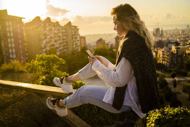 Young woman sitting on railing above the city using cell phone, Barcelona, Spain - GIOF07950