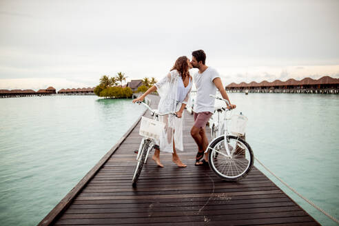 Pärchen mit Fahrrädern küsst sich auf einem Steg im Meer, Insel Maguhdhuvaa, Gaafu Dhaalu Atoll, Malediven - DAWF01235
