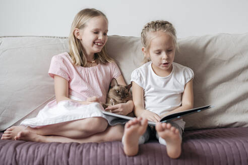 Two little girls sitting barefoot on the couch with Burmese cat watching a book - EYAF00914