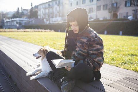 Junge Frau mit Piercings und Zöpfen sitzt mit ihrem Hund auf einer Bank und liest ein Buch, Como, Italien, lizenzfreies Stockfoto