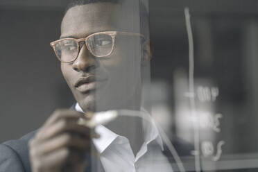Portrait of content young businessman drawing diagram on glass pane - KNSF07415