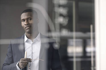 Portrait of smiling young businessman behind diagram on glass pane in office - KNSF07417