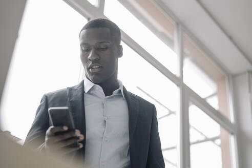 Portrait of young businessman standing in front of window looking at cell phone - KNSF07422