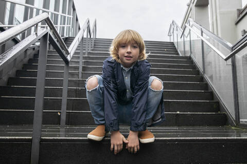 Porträt eines blonden Jungen, der auf einer Treppe im Freien hockt, lizenzfreies Stockfoto