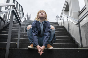 Porträt eines blonden Jungen, der auf einer Treppe im Freien kauert und nach oben schaut - EYAF00924