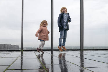 Bruder und kleine Schwester springen auf der Aussichtsterrasse an einem regnerischen Tag - EYAF00926