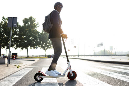 Businessman with backpack and cycling helmet crossing street on push scooter at sunset - JSRF00783