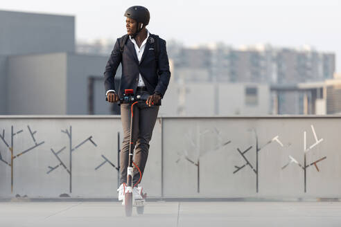 Businessman with backpack and cycling helmet riding push scooter in the evening - JSRF00794