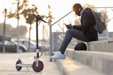 Junger Geschäftsmann mit Tretroller, der bei Sonnenuntergang auf einer Treppe sitzt und Mobiltelefon und Kopfhörer benutzt - JSRF00795