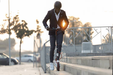 Young businessman with backpack and cycling helmet riding push scooter at sunset - JSRF00796