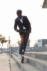 Portrait of businessman with backpack and cycling helmet riding push scooter at sunset - JSRF00797