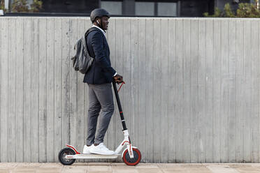 Smiling businessman on push scooter in front of concrete wall - JSRF00798