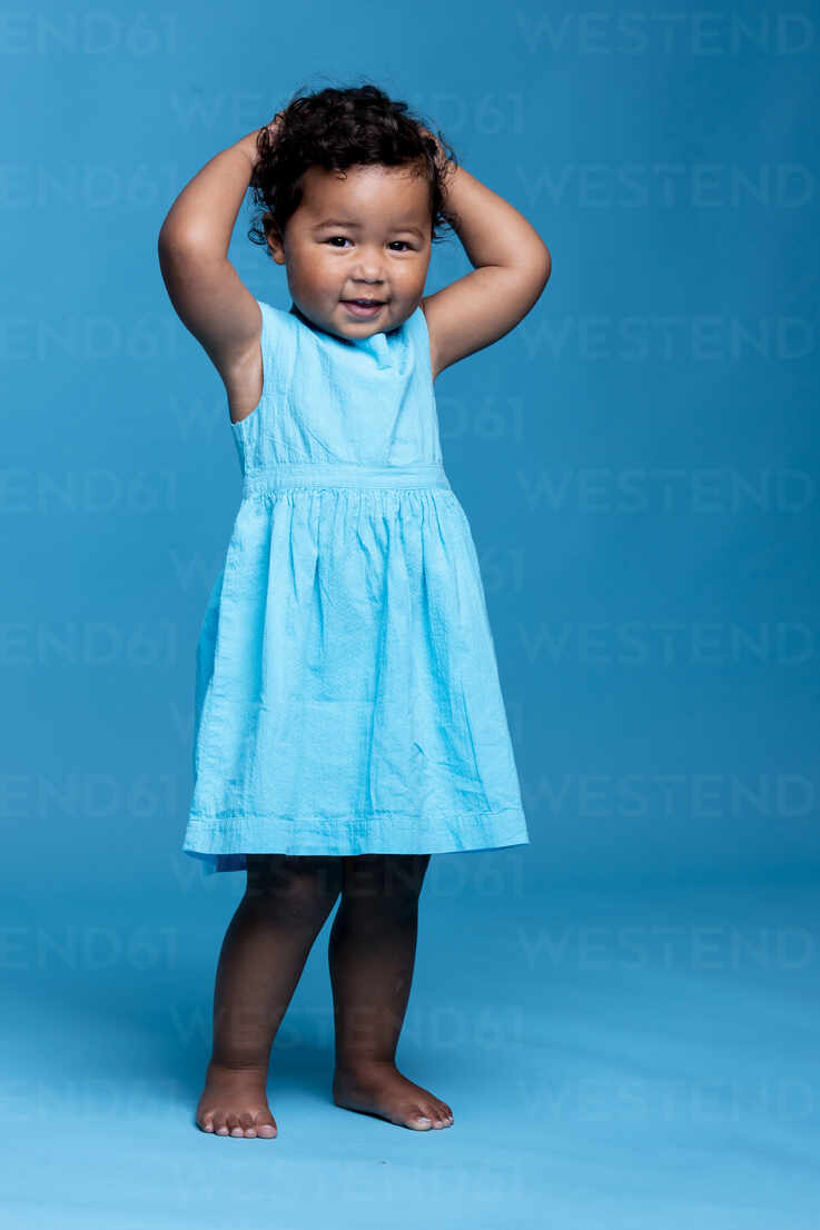 Portrait of smiling little girl with hands on head wearing light blue dress  standing against blue background stock photo