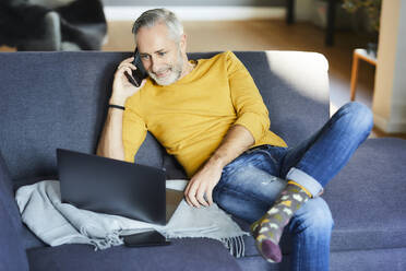 Mature man using cell phone and laptop on couch at home - FMKF06120