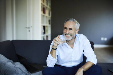 Lächelnder reifer Mann trinkt ein Glas Wasser auf dem Sofa zu Hause - FMKF06127