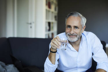 Lächelnder reifer Mann trinkt ein Glas Wasser auf dem Sofa zu Hause - FMKF06128
