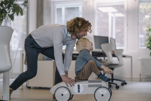 Businessman pushing son on toy car in office - KNSF07466