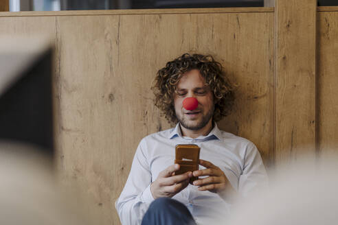 Geschäftsmann mit roter Clownsnase benutzt Handy im Büro - KNSF07503