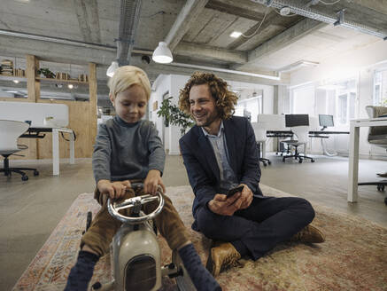 Smiling businessman with son on toy car in office - KNSF07519