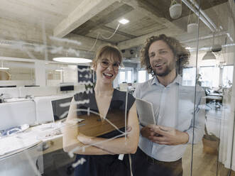 Porträt eines glücklichen Geschäftsmannes und einer Geschäftsfrau hinter einer Glasscheibe im Büro - KNSF07520