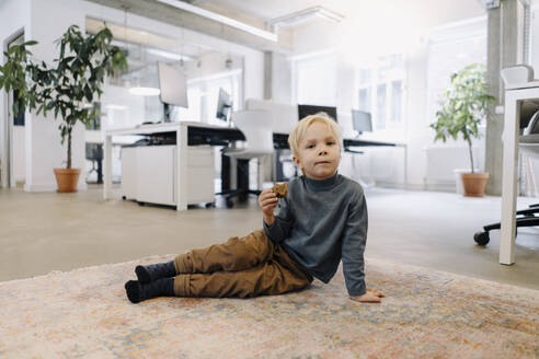 Junge sitzt auf Teppich im Büro und isst Brot - KNSF07533