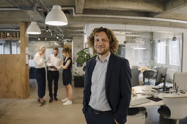 Portrait of confident businessman in office with colleagues in background - KNSF07545