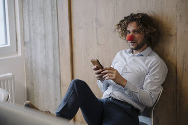 Porträt eines Geschäftsmannes mit roter Clownsnase, der im Büro ein Mobiltelefon benutzt - KNSF07591