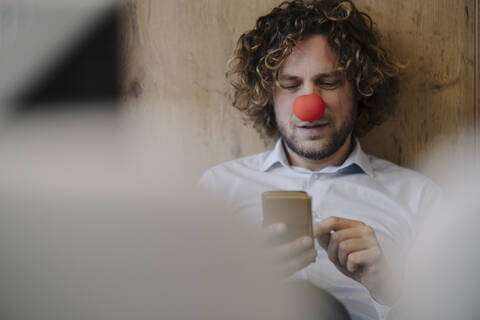 Geschäftsmann mit roter Clownsnase benutzt Handy im Büro, lizenzfreies Stockfoto