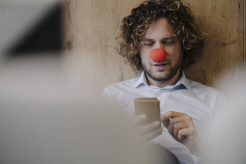 Geschäftsmann mit roter Clownsnase benutzt Handy im Büro - KNSF07592