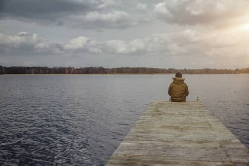 Rückenansicht eines jungen Mannes, der auf einem Steg sitzt und auf einen See im Winter schaut - ANHF00155