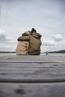 Rückenansicht eines jungen verliebten Paares, das auf einem Steg sitzt und auf einen See im Winter blickt - ANHF00156