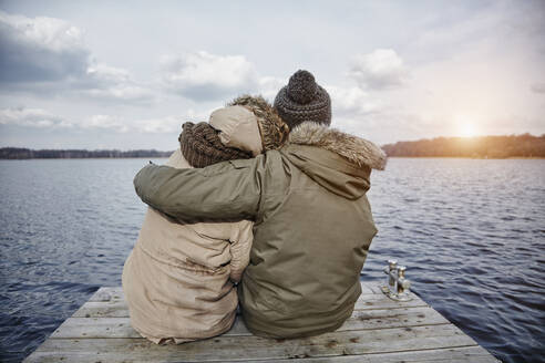 Rückenansicht eines jungen verliebten Paares, das auf einem Steg sitzt und auf einen See im Winter blickt - ANHF00157