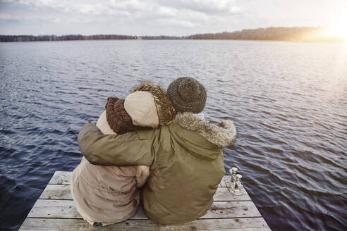 Rückenansicht eines jungen verliebten Paares, das auf einem Steg sitzt und auf einen See im Winter blickt - ANHF00158