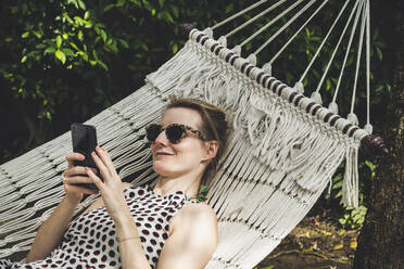 Lächelnde Frau in einer Hängematte liegend, die ein Mobiltelefon benutzt - CHPF00602