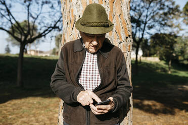 Alter Mann mit Smartphone, an Baumstamm gelehnt - JRFF04105