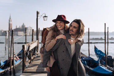 Young man giving his girlfriend a piggyback ride at the waterfront in Venice, Italy - MAUF03264