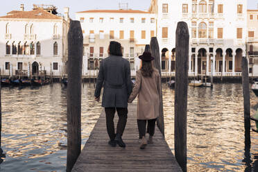 Rückansicht eines jungen Paares, das auf einem Steg am Canal Grande in Venedig, Italien, spazieren geht - MAUF03294