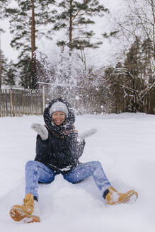 Glückliche Frau sitzt auf einem Schneefeld und spielt mit Schneebällen - KNTF04190